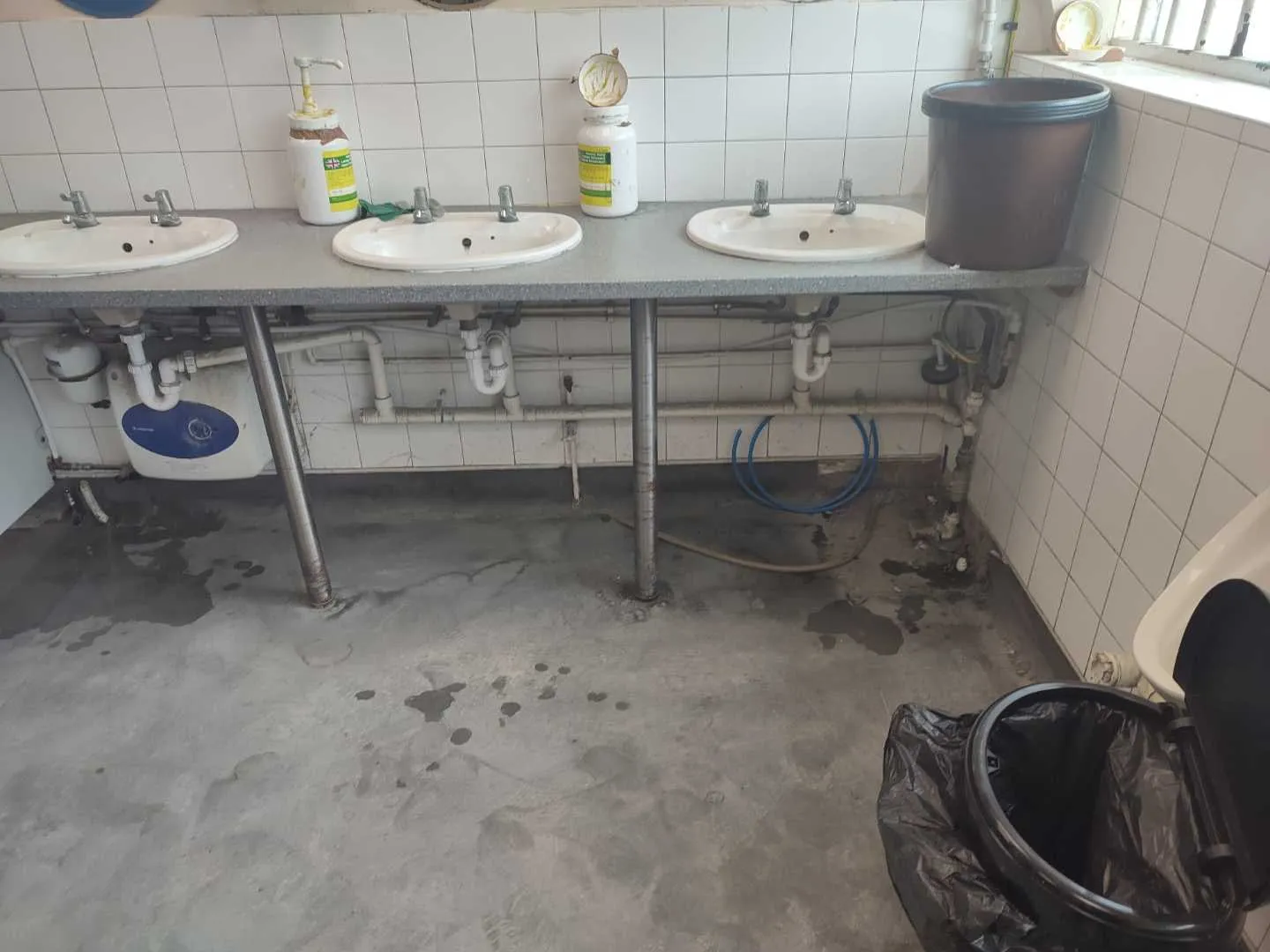 Factory washroom with three basins on a counter and exposed pipework and a water heater beneath it